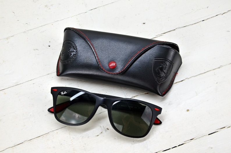 K&ouml;nnte beinhalten: Schwarze Sonnenbrille mit roten Akzenten und einem schwarzen Lederetui mit roten N&auml;hten. Das Etui hat ein gepr&auml;gtes Ferrari-Logo.