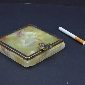 Può includere: Un portacenere quadrato in marmo verde e bianco con un bordo in metallo argentato e un design a farfalla sulla parte superiore. Una sigaretta bianca giace su una superficie nera accanto al portacenere.