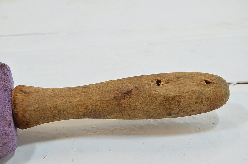 Puede incluir: Un rodillo de madera con una esponja morada unida a un extremo. El rodillo tiene dos agujeros perforados en el mango.