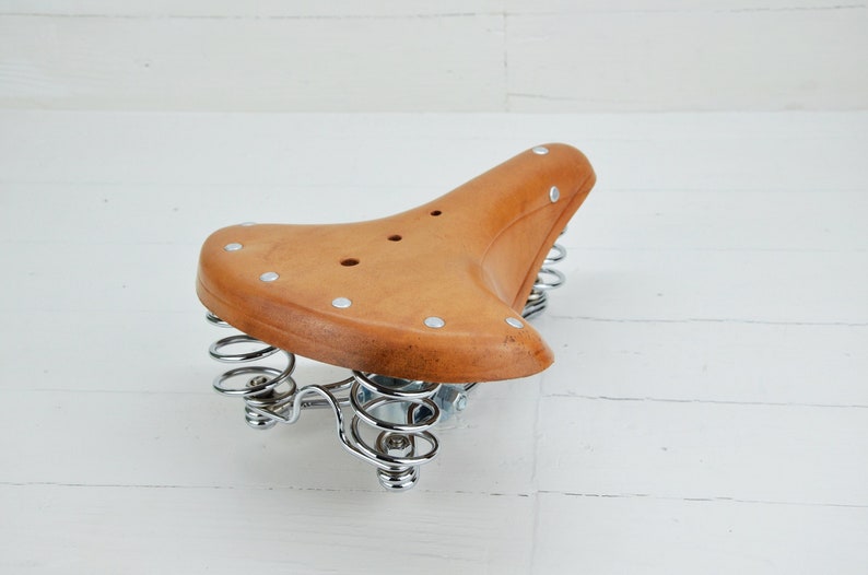 May include: A vintage-style bicycle seat in light brown leather. The seat has a curved shape with silver rivets and is supported by two chrome-plated springs. The seat is designed for comfort and style.