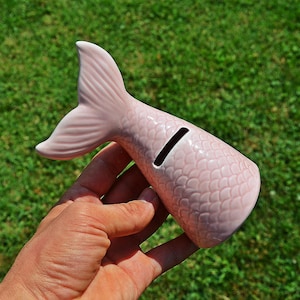 Può includere: Salvadanaio in ceramica rosa a forma di coda di sirena con uno slot per le monete. La coda è testurizzata per sembrare squame.