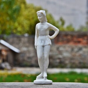 以下が含まれることがあります： 腰に手を当てて立っている女性の白い磁器の置物。置物はシンプルなドレスを着ており、顔は中立的な表情をしている。