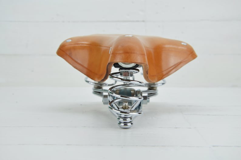 May include: A brown leather bicycle saddle with a chrome-plated spring suspension system. The saddle is designed for comfort and features a classic, vintage aesthetic. The springs provide shock absorption for a smoother ride.
