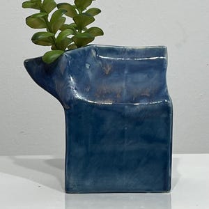 Può includere: Un vaso in ceramica blu con una finitura lucida e irregolare e un corpo rettangolare caratterizzato da un beccuccio angolato sul lato sinistro. Una piccola pianta succulenta verde con foglie arrotondate è inserita nell'apertura del vaso.