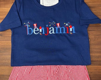 May include: A navy blue t-shirt with a red, white, and blue embroidered design that says "Benjamin" with fireworks and top hats. The shirt is paired with red and white gingham shorts.