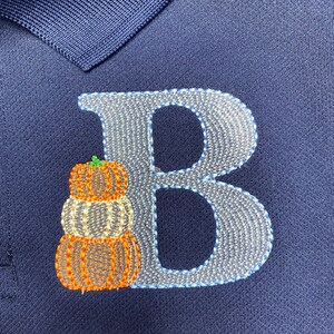 Puede incluir: Una camiseta polo azul marino con una letra "B" bordada en blanco y una pila de tres calabazas naranjas.