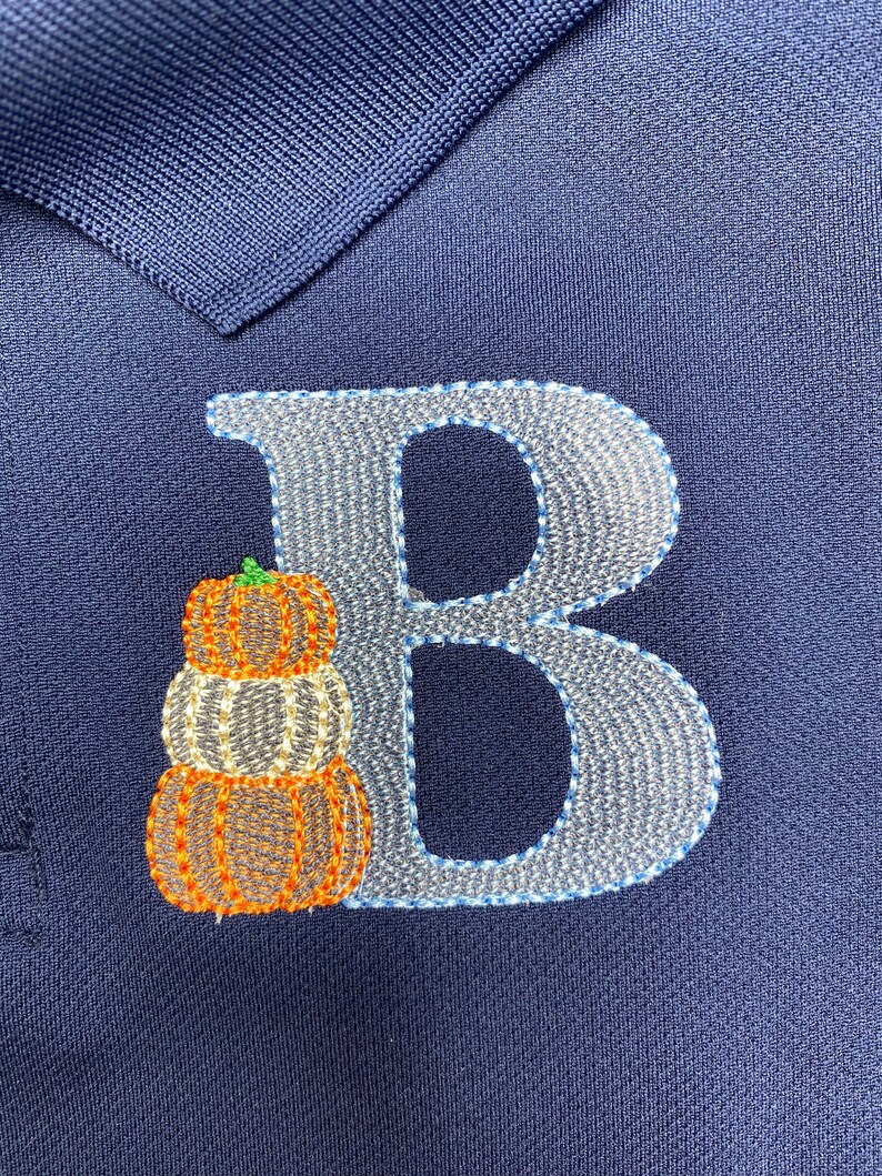 Puede incluir: Una camiseta polo azul marino con una letra "B" bordada en blanco y una pila de tres calabazas naranjas.