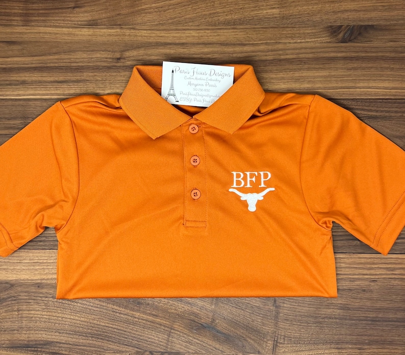 May include: Orange polo shirt with a white embroidered logo that says "BFP" and a white Longhorn steer head. The shirt is on a wooden surface.