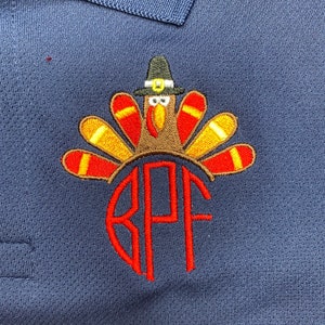 Puede incluir: Una camisa polo azul marino con un dise&ntilde;o de pavo de Acci&oacute;n de Gracias bordado. El pavo lleva un sombrero de peregrino y tiene un dise&ntilde;o de plumas rojas, amarillas y marrones. La camisa tambi&eacute;n tiene un monograma "RPF" bordado en rojo.