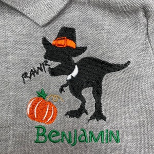 Puede incluir: Una camiseta polo gris con un dise&ntilde;o bordado de un dinosaurio negro que lleva un sombrero de peregrino y sostiene una calabaza. El dinosaurio dice "RAWR" y la camiseta dice "Benjamin".