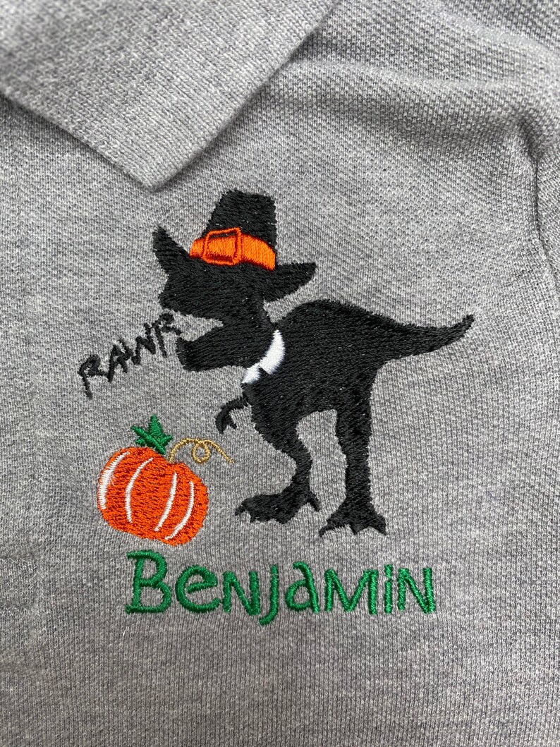 Puede incluir: Una camiseta polo gris con un dise&ntilde;o bordado de un dinosaurio negro que lleva un sombrero de peregrino y sostiene una calabaza. El dinosaurio dice "RAWR" y la camiseta dice "Benjamin".