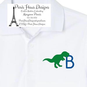 May include: White polo shirt with a green dinosaur and blue letter B embroidered on the front. The shirt has a small embroidered logo with the text "Paris Haus Designs, Custom Machine Embroidery, Maryann Parris, 512-736-1830, Paris Haus Designs@gmail.com, ETSY: Paris Haus Designs".