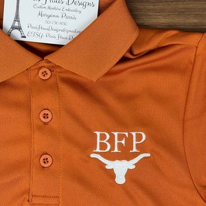 May include: A burnt orange polo shirt with white embroidery of the letters "BFP" and a white stylized Longhorn steer head. The shirt has a button-up collar and three buttons down the front.