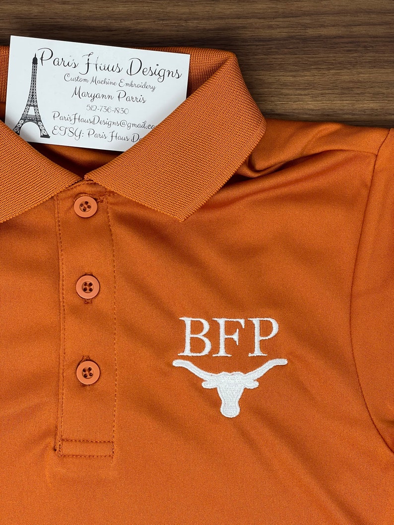 May include: A burnt orange polo shirt with white embroidery of the letters "BFP" and a white stylized Longhorn steer head. The shirt has a button-up collar and three buttons down the front.