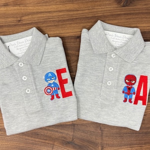 May include: Two gray polo shirts with embroidered superhero characters and letters. The left shirt features Captain America with the letter "E" and the right shirt features Spiderman with the letter "A".