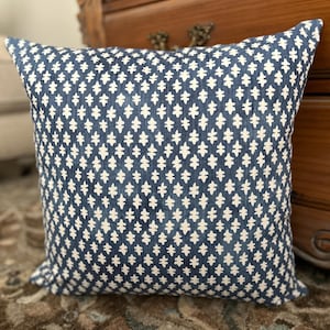 May include: A square throw pillow featuring a deep blue fabric base with a repeating white geometric pattern of small, stylized cross-like shapes. The textile has a visible woven texture, and the design is consistent across the entire front surface.