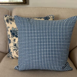 May include: A square throw pillow featuring a blue and white gingham check pattern on the front. Behind it sits a second square throw pillow with a cream-colored background and a blue floral pattern. Both pillows are made of fabric.