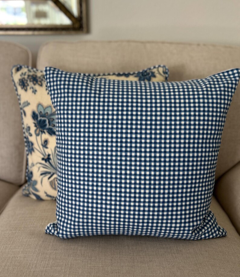 May include: A square throw pillow featuring a blue and white gingham check pattern on the front. Behind it sits a second square throw pillow with a cream-colored background and a blue floral pattern. Both pillows are made of fabric.