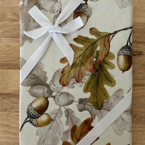 May include: A rectangular, fabric-covered item with a fall-themed acorn and oak leaf pattern. The item is wrapped with a white ribbon tied in a bow. The background is a light beige color.