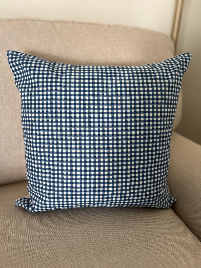 May include: A square throw pillow featuring a classic navy blue and white gingham check pattern. The fabric displays a consistent grid of small, alternating dark blue and white squares across the entire surface of the cushion cover.