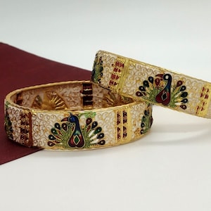 May include: Two white bangle bracelets with gold accents and peacock designs. The peacocks are decorated with blue, green, red, and gold enamel.