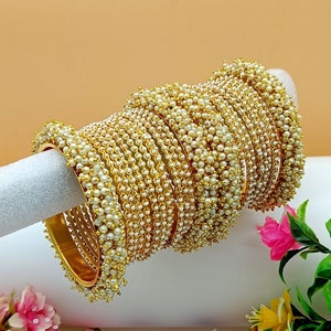 May include: A stack of gold bangles with white pearl accents. The bangles are arranged in a circular pattern and are displayed on a white surface.