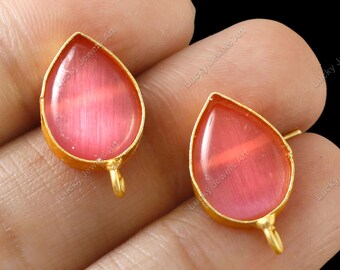 May include: A pair of teardrop-shaped earrings with a gold-colored frame. The earrings feature a pink, translucent stone. Each earring has a small loop at the bottom. The background is black.