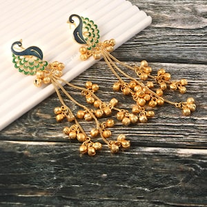 May include: A pair of gold-colored earrings featuring peacock designs. The earrings have a peacock head with blue and green enamel detailing, and dangling chains with small bell charms. The earrings are displayed on a white surface.