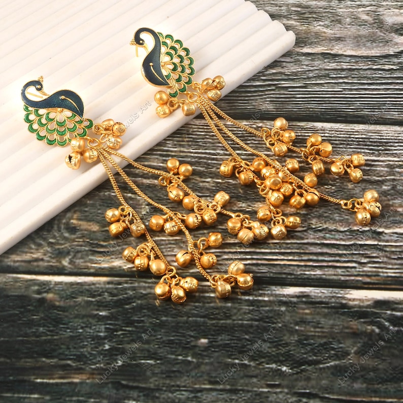 May include: A pair of gold-colored earrings featuring peacock designs. The earrings have a peacock head with blue and green enamel detailing, and dangling chains with small bell charms. The earrings are displayed on a white surface.