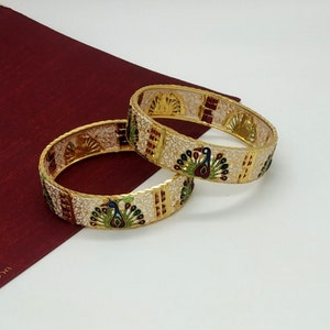 May include: Two gold bangles with white enamel and peacock designs. The peacocks are decorated with green, blue, and red enamel.