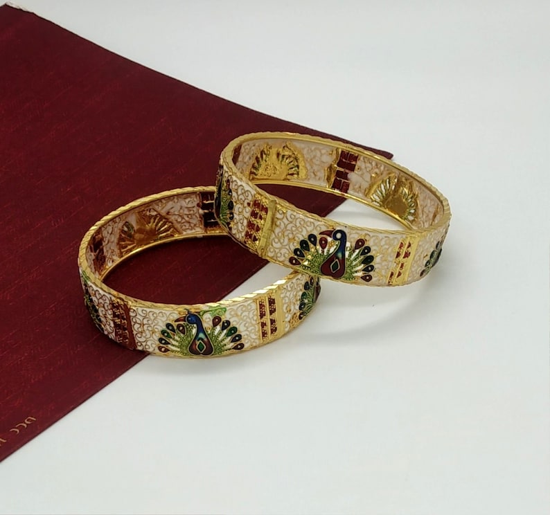 May include: Two gold bangles with white enamel and peacock designs. The peacocks are decorated with green, blue, and red enamel.