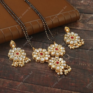 May include: A gold necklace with three flower-shaped pendants and a red stone in the center of each flower. The necklace is on a black string and has white pearl accents. There are two matching earrings with the same design.