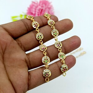 Op de afbeelding: Twee gouden armbanden met ronde, sierlijke bedels. Elke bedel heeft een centrale steen, een groene en een roze, omringd door kleinere witte stenen.