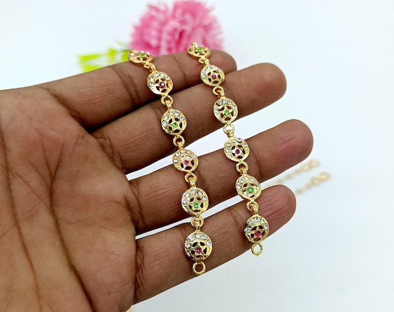 Op de afbeelding: Twee gouden armbanden met ronde, sierlijke bedels. Elke bedel heeft een centrale steen, een groene en een roze, omringd door kleinere witte stenen.
