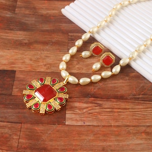 May include: A gold-toned pendant necklace and matching earrings set. The pendant features a large red gemstone surrounded by smaller red and green accents. The earrings and necklace are adorned with faux pearls.