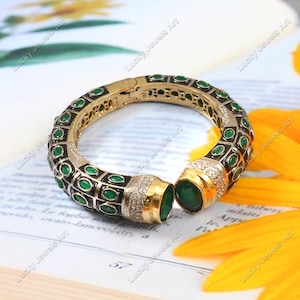 May include: A gold bangle bracelet with a black and green enamel design. The bracelet is decorated with small green gemstones and clear rhinestones.