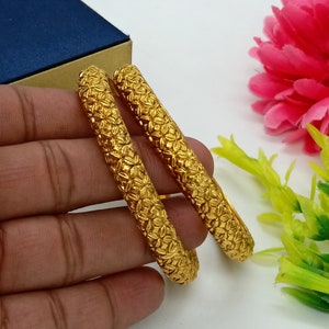 以下が含まれることがあります： 花柄の金色のバングル2つ。バングルは重ねられています。