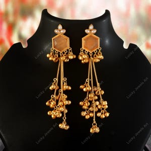 May include: A pair of gold-colored dangle earrings. Each earring features a hexagonal top with a peach-colored stone and small white accents. Below, chains cascade with clusters of small, round beads. The earrings are displayed on a black stand.