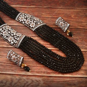 May include: A black beaded necklace and matching earrings set. The necklace has multiple strands of black beads, with silver-toned clasps decorated with white floral designs. The earrings have a similar design, with a black bead drop.