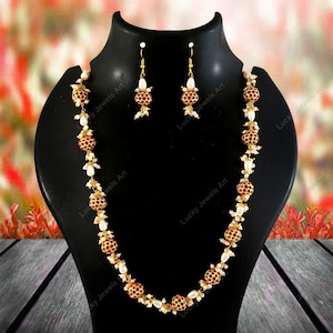 May include: A gold-toned necklace and earring set. The necklace features alternating red and gold beaded spheres and white pearl-like accents. The earrings mirror the design with a single red and gold sphere and pearl accents. The jewelry is displayed on a black mannequin.