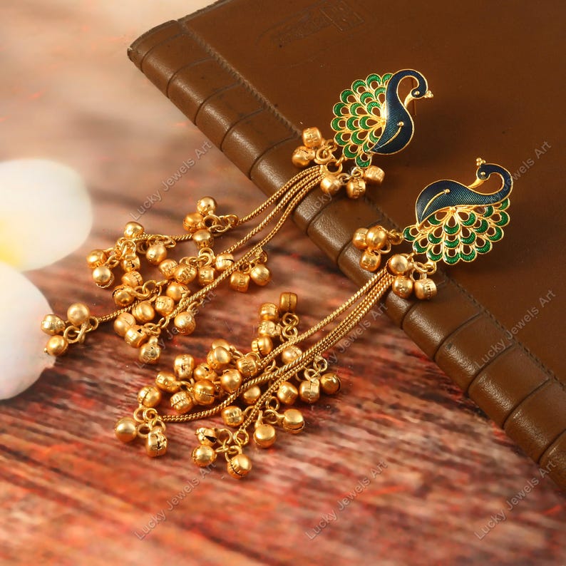 May include: Gold-toned peacock earrings with blue and green enamel detailing. The earrings feature a dangling chain with small, round gold beads. The peacock design is intricate, with a detailed feather pattern. The earrings are displayed on a brown surface.