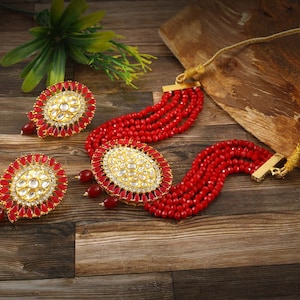 May include: A red beaded necklace and matching earrings set. The necklace features multiple strands of red beads and a central oval pendant with a gold-colored frame and white accents. The earrings have a similar design, with red teardrop-shaped accents.