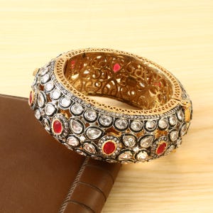 Op de afbeelding: Een gouden armband met ingewikkelde details en een zwarte emaille afwerking. De armband is versierd met witte en rode edelstenen.