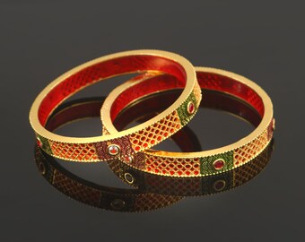 May include: Two gold bangles with detailed patterns. The bangles have a red interior and are decorated with red, green, and maroon accents. The design includes a lattice pattern and small, round gemstones. The bangles are displayed on a reflective surface.