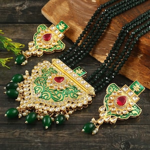 Può includere: Un set di collana e orecchini dorati con dettagli in smalto verde e accenti di pietre preziose rosse. La collana presenta più fili di perline verde scuro. Gli orecchini e il ciondolo hanno disegni intricati e perline verdi.