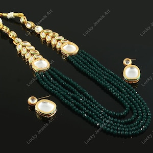 Op de afbeelding: Een meerrijige ketting met groene kralen en gouden accenten. De ketting heeft een hanger met een grote, heldere, ronde steen omgeven door kleinere stenen. De ketting wordt geleverd met bijpassende oorbellen.