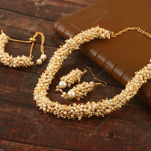 Puede incluir: Un conjunto de collar, pulsera y pendientes de oro con cuentas de perlas blancas. Las joyas están sobre una superficie de madera marrón.