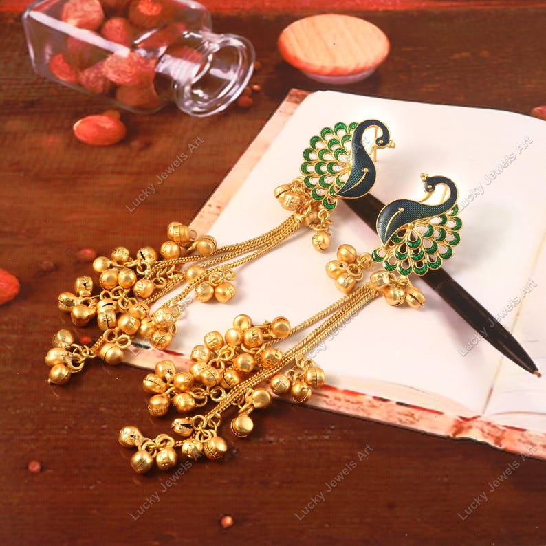 May include: A pair of gold-colored earrings with a peacock design. The earrings feature a peacock with a blue body and green tail feathers. Dangling from the peacock are gold chains with small, round gold bells. The earrings are on a white surface.