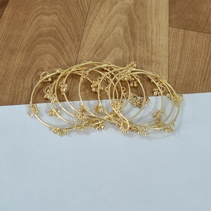 May include: A set of gold-coloured bangles, each adorned with tiny, bell-shaped dangles. The bangles are arranged in a slightly overlapping manner, highlighting their intricate design. The background features light wood and white paper.