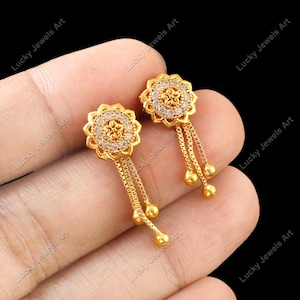 Könnte beinhalten: Ein Paar goldfarbene Ohrringe mit floralem Design. Jeder Ohrring hat eine blütenförmige Oberseite mit kleinen, klaren Steinen und herabhängenden Ketten, die in kleinen goldenen Perlen enden. Die Ohrringe werden in einer Hand vor schwarzem Hintergrund gehalten.